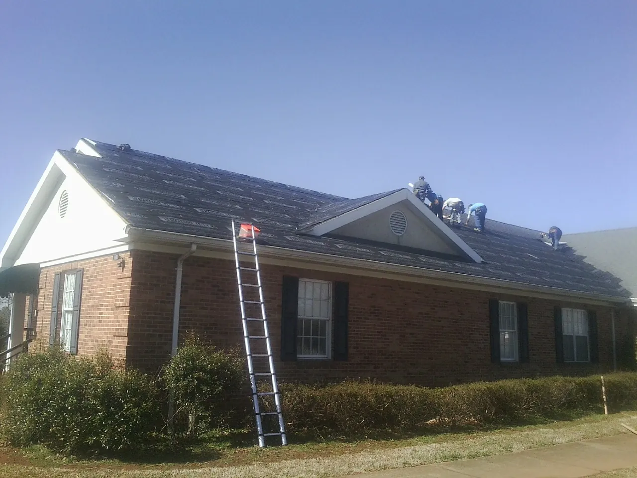 Crew tearing shingles off a brick ranch roof with a ladder set — Emergency Roof Repair work by our crew in Bennettsville