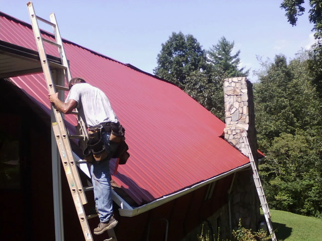 Roofer on a ladder beside a red metal roof and stone chimney — Soffit & Fascia Repair by Roofing Brothers in Bennettsville
